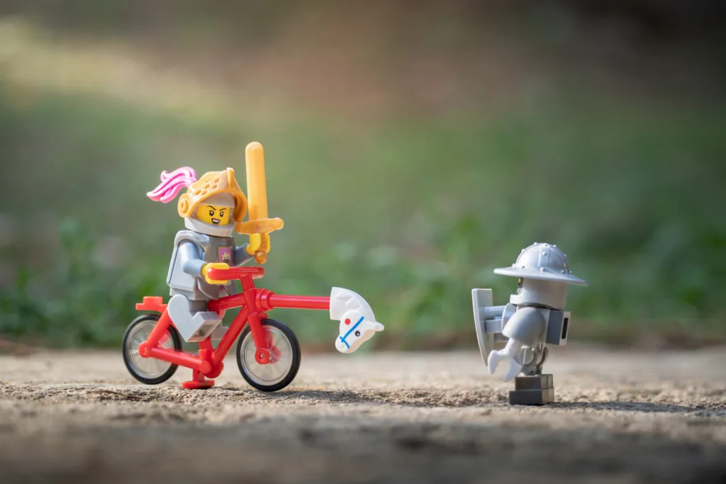 Photo of LEGO knight on a bicycle with a hobby horse attached charging at a robot guard